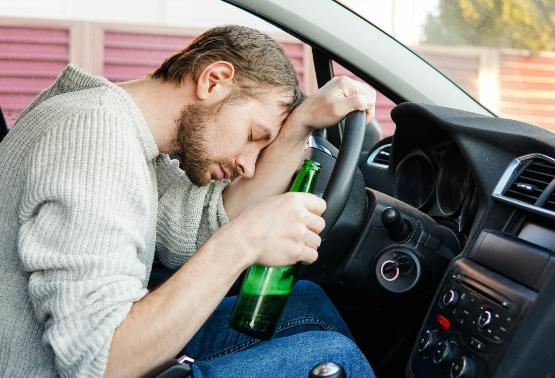 A man slumped over a car steering wheel holding a beer bottle, illustrating the dangers of drink-driving and its impact on motor trade insurance.